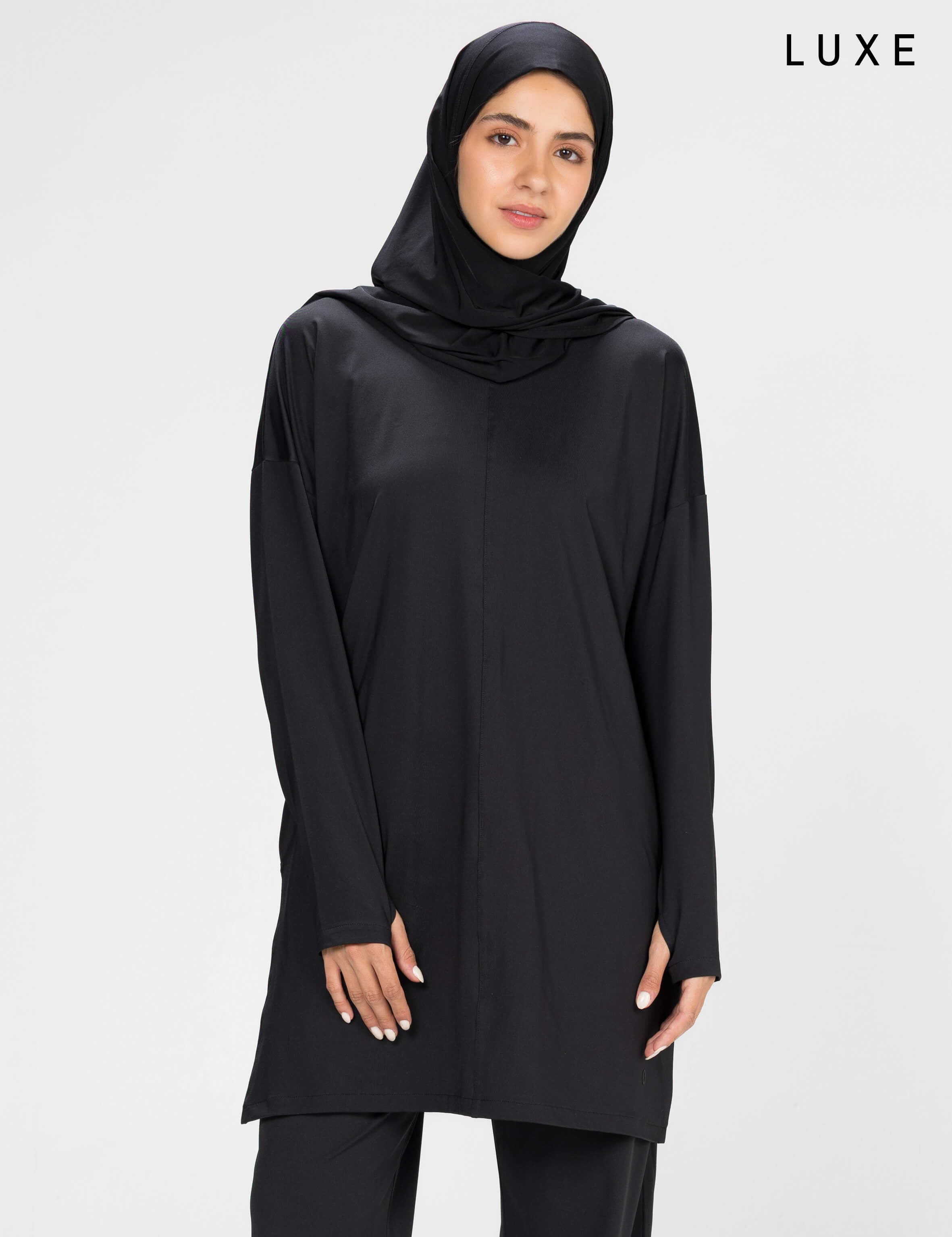 Woman wearing a long-sleeve black modest loose sports activewear top with a hijab on a white background, branded 'LUXE'.