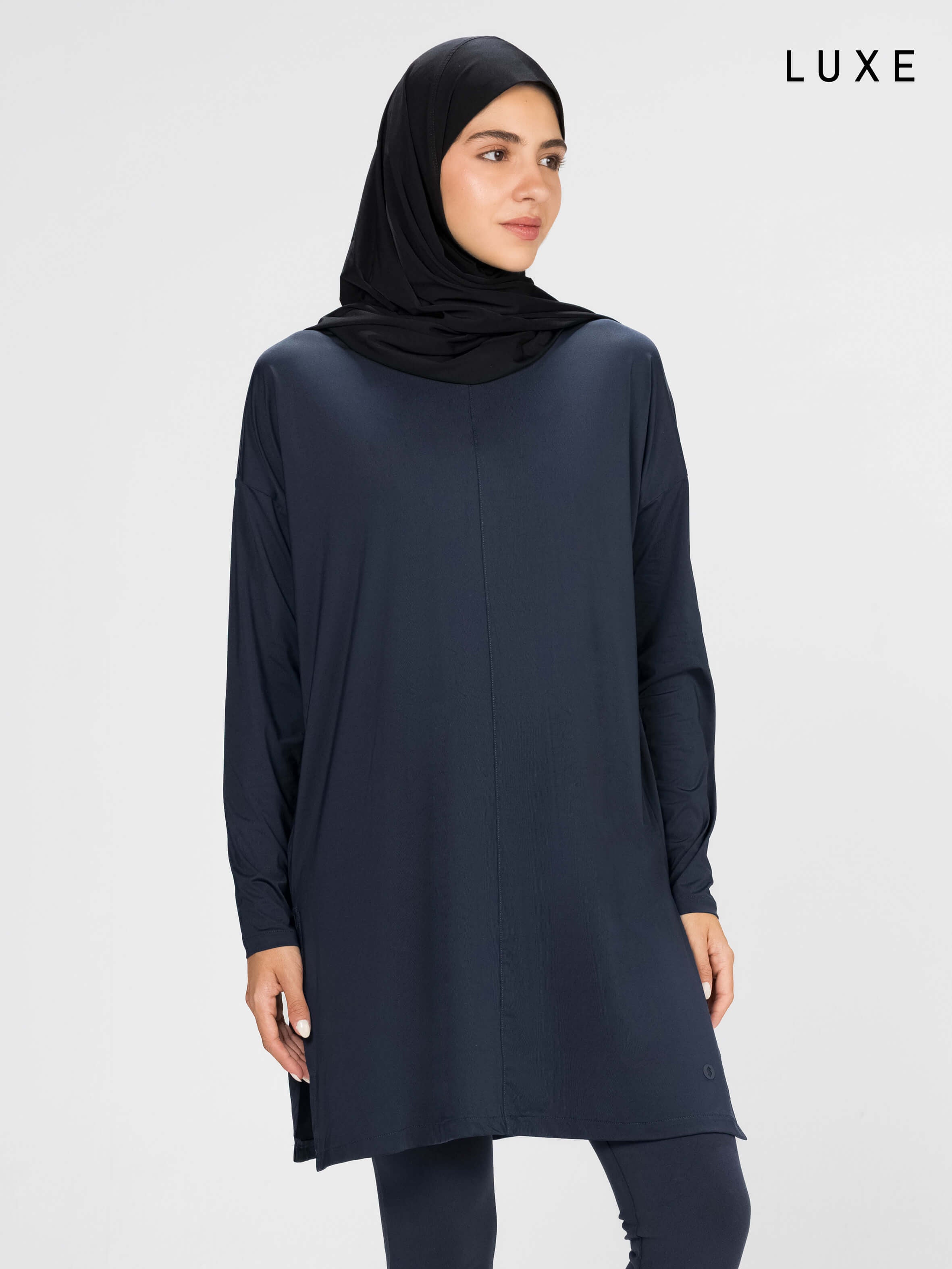 Woman wearing a long-sleeve navy modest loose sports activewear top with a hijab on a white background, branded 'LUXE'.