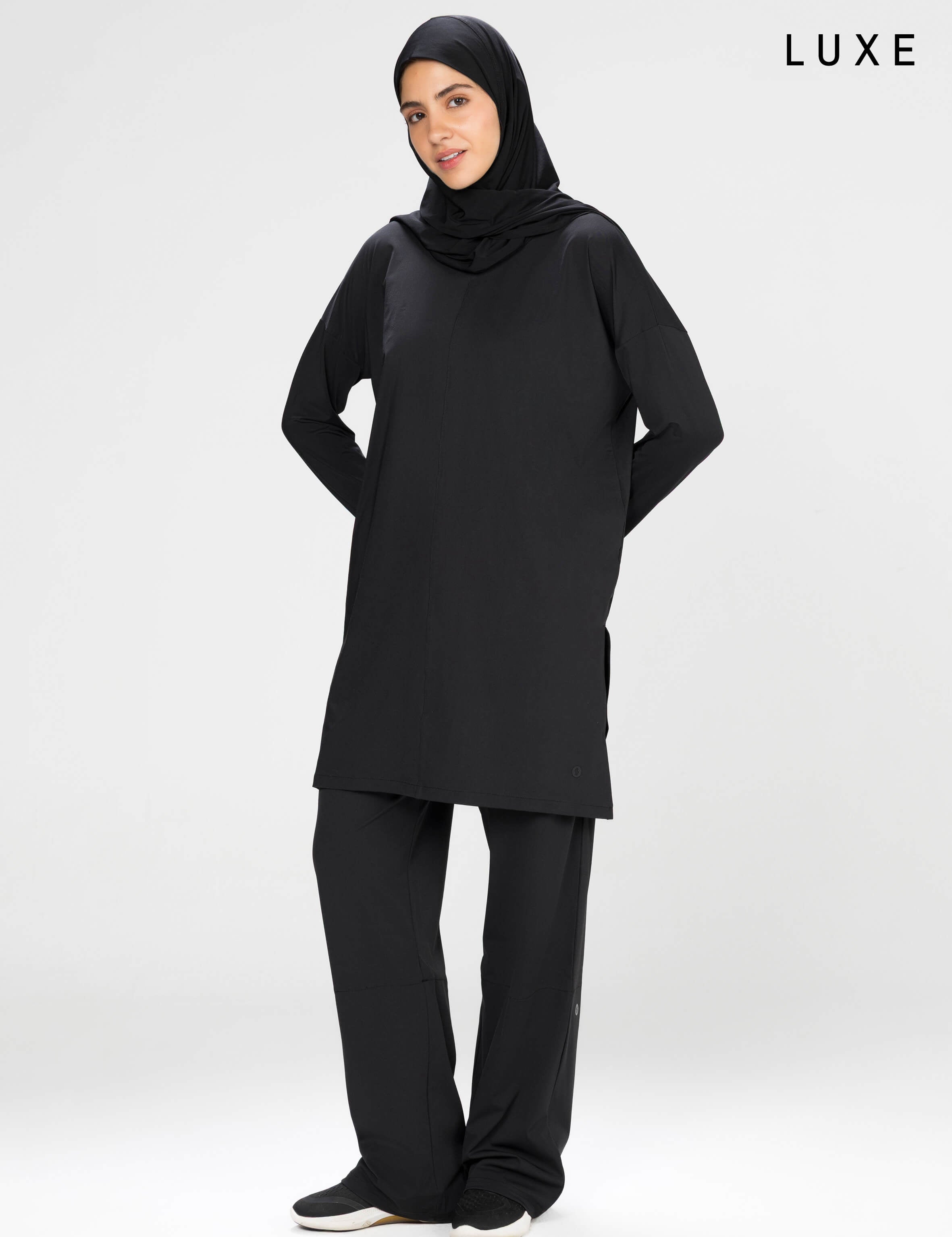 Woman wearing a long-sleeve black modest loose sports activewear top with a hijab on a white background, branded 'LUXE'.