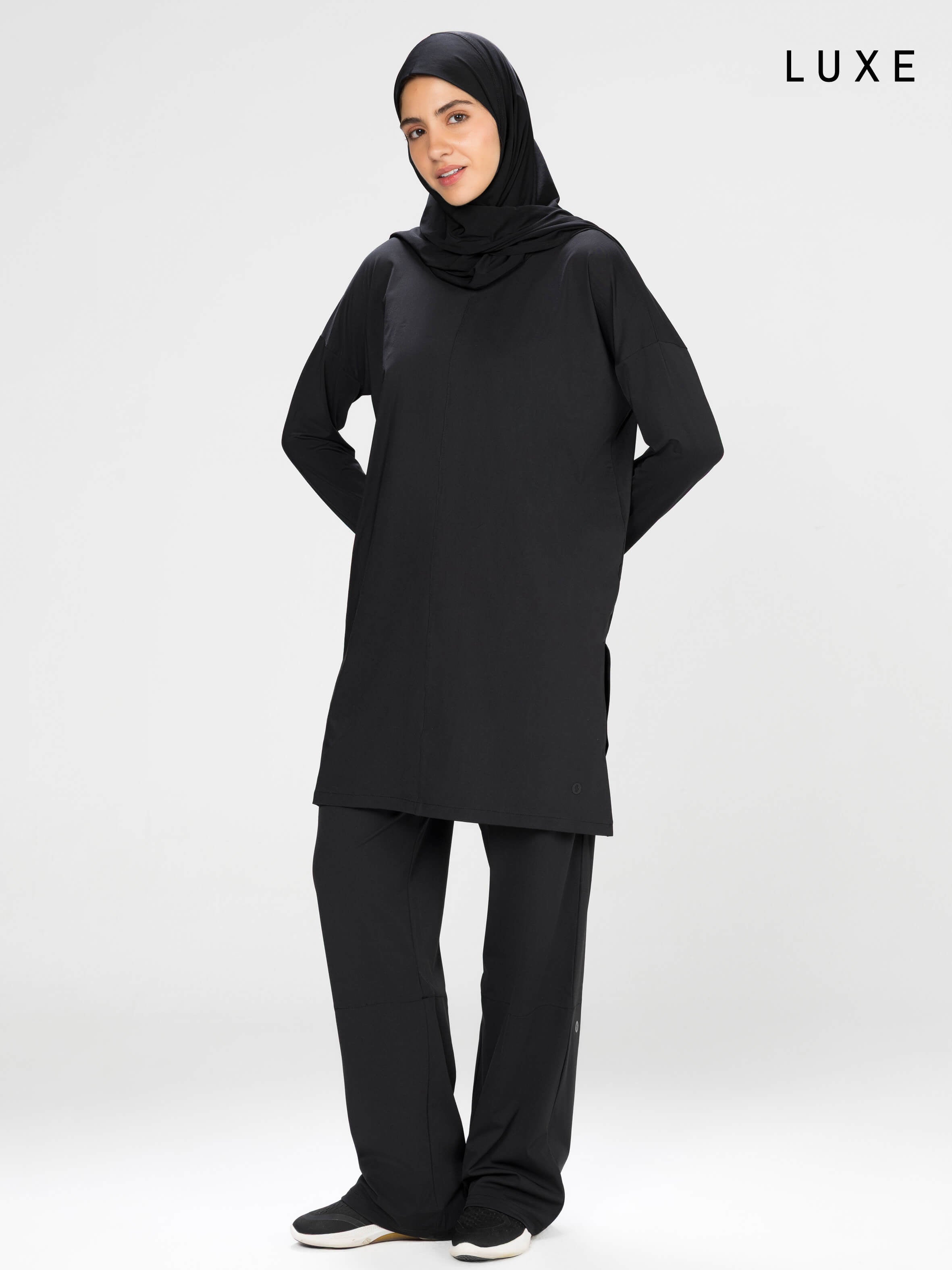 Woman wearing a long-sleeve black modest loose sports activewear top with a hijab on a white background, branded 'LUXE'.