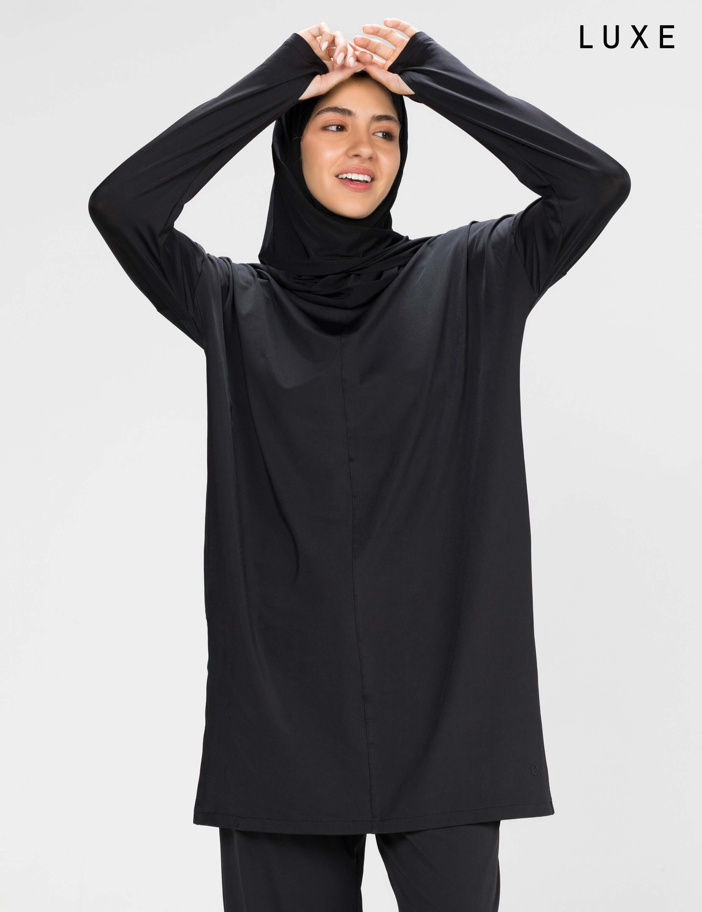 Woman wearing a long-sleeve black modest loose sports activewear top with a hijab on a white background, branded 'LUXE'.