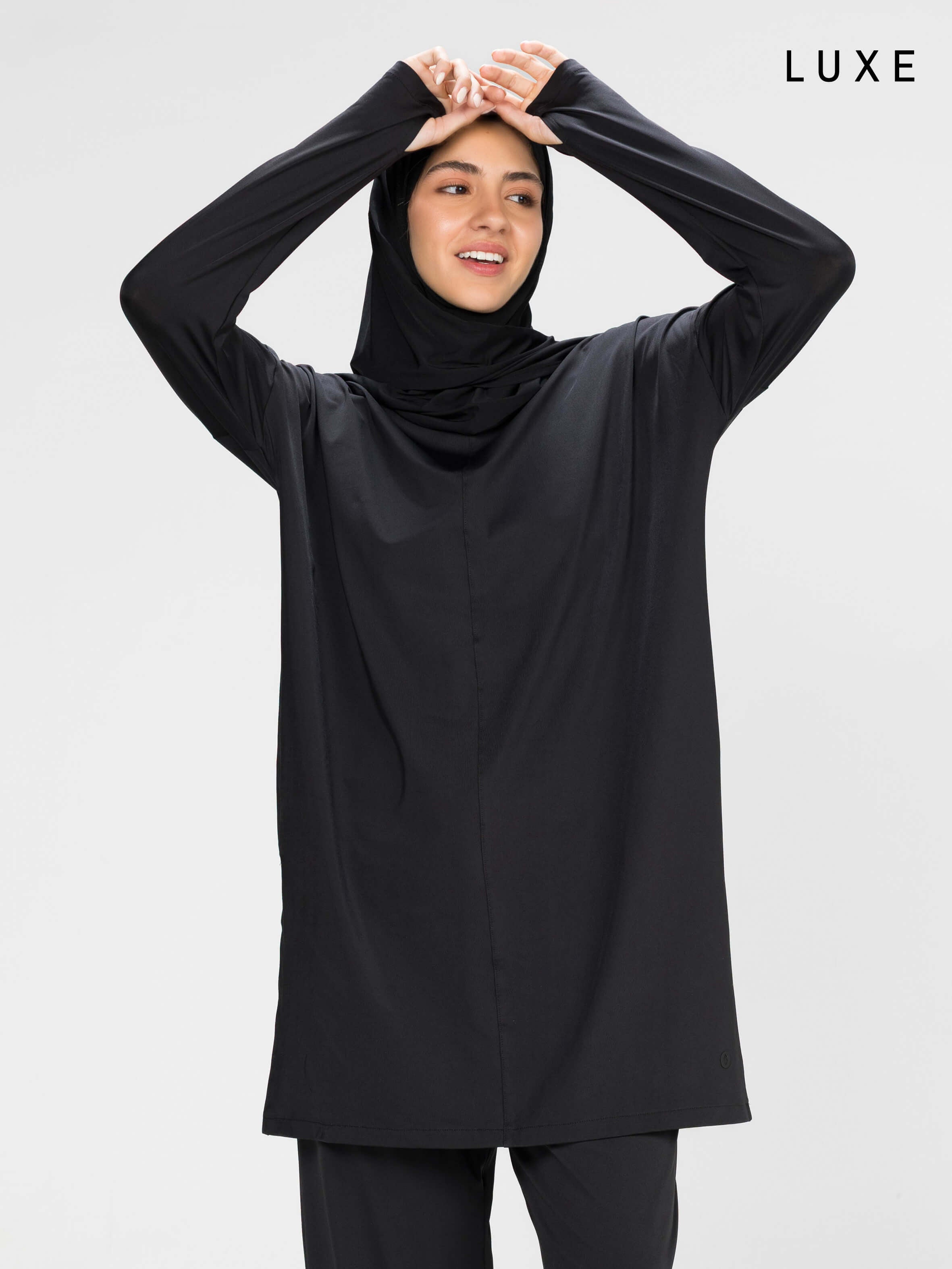 Woman wearing a long-sleeve black modest loose sports activewear top with a hijab on a white background, branded 'LUXE'.