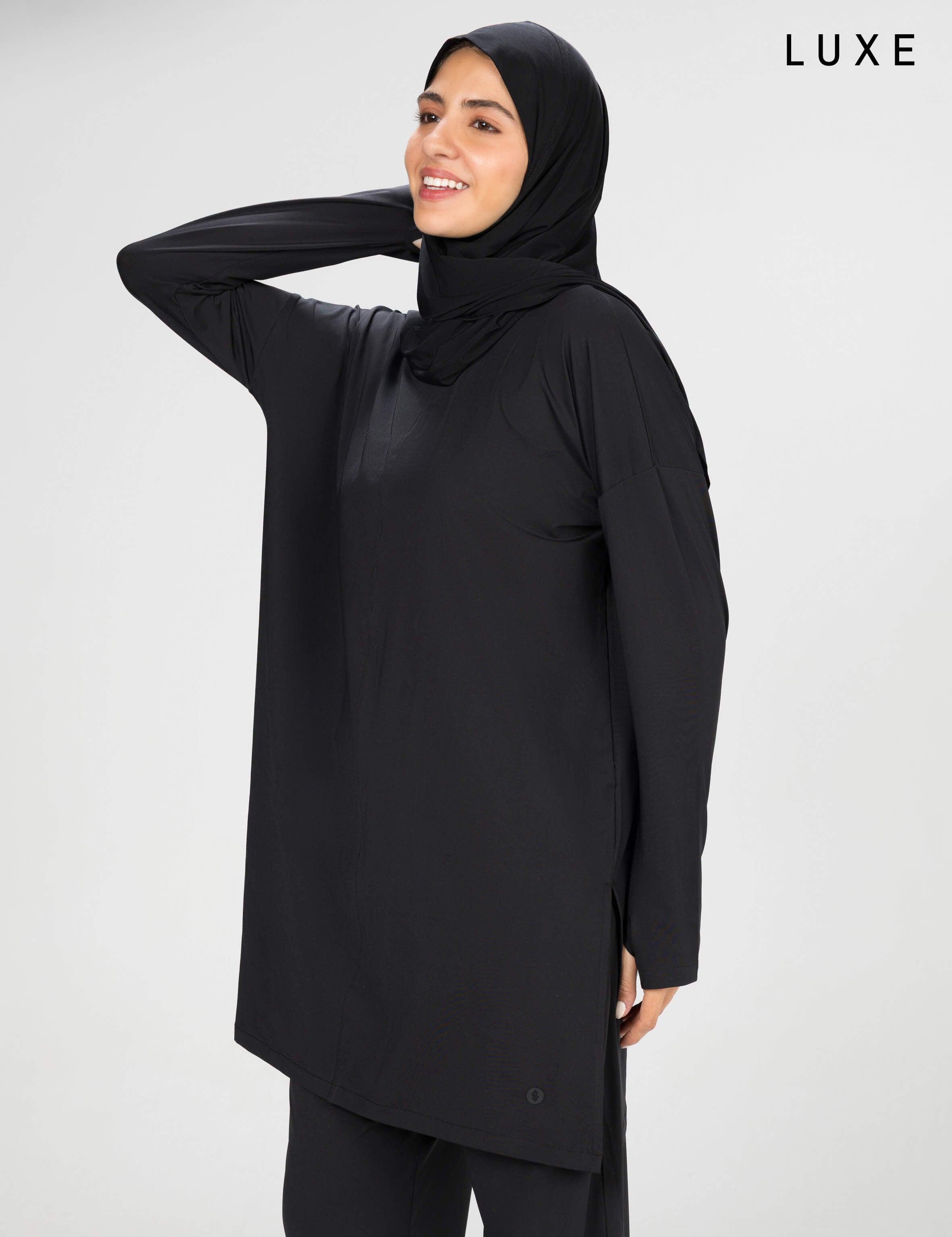 Woman wearing a long-sleeve black modest loose sports activewear top with a hijab on a white background, branded 'LUXE'.