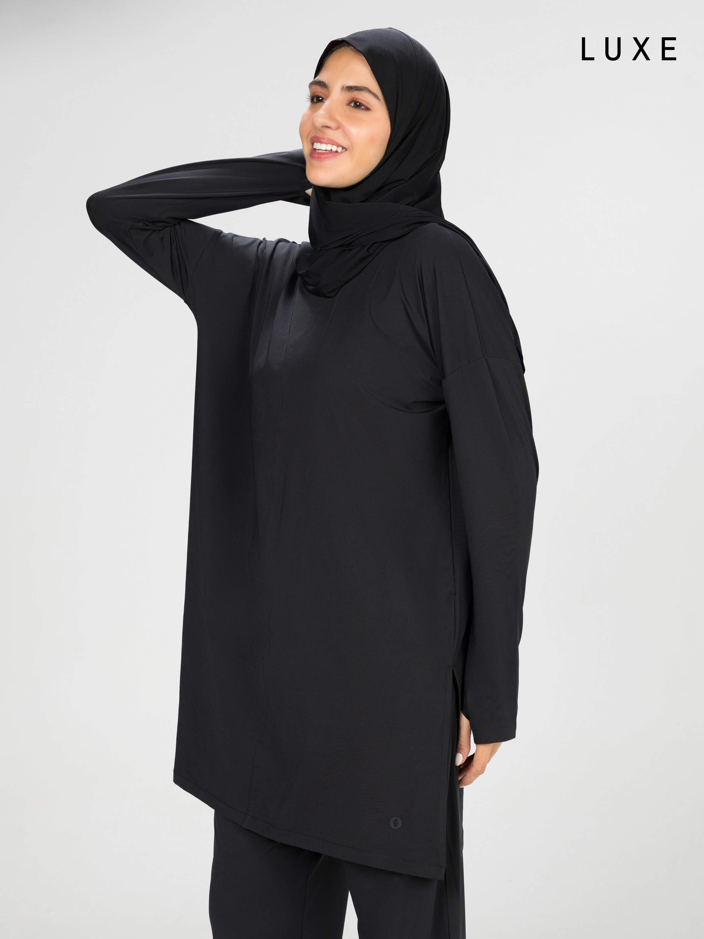 Woman wearing a long-sleeve black modest loose sports activewear top with a hijab on a white background, branded 'LUXE'.