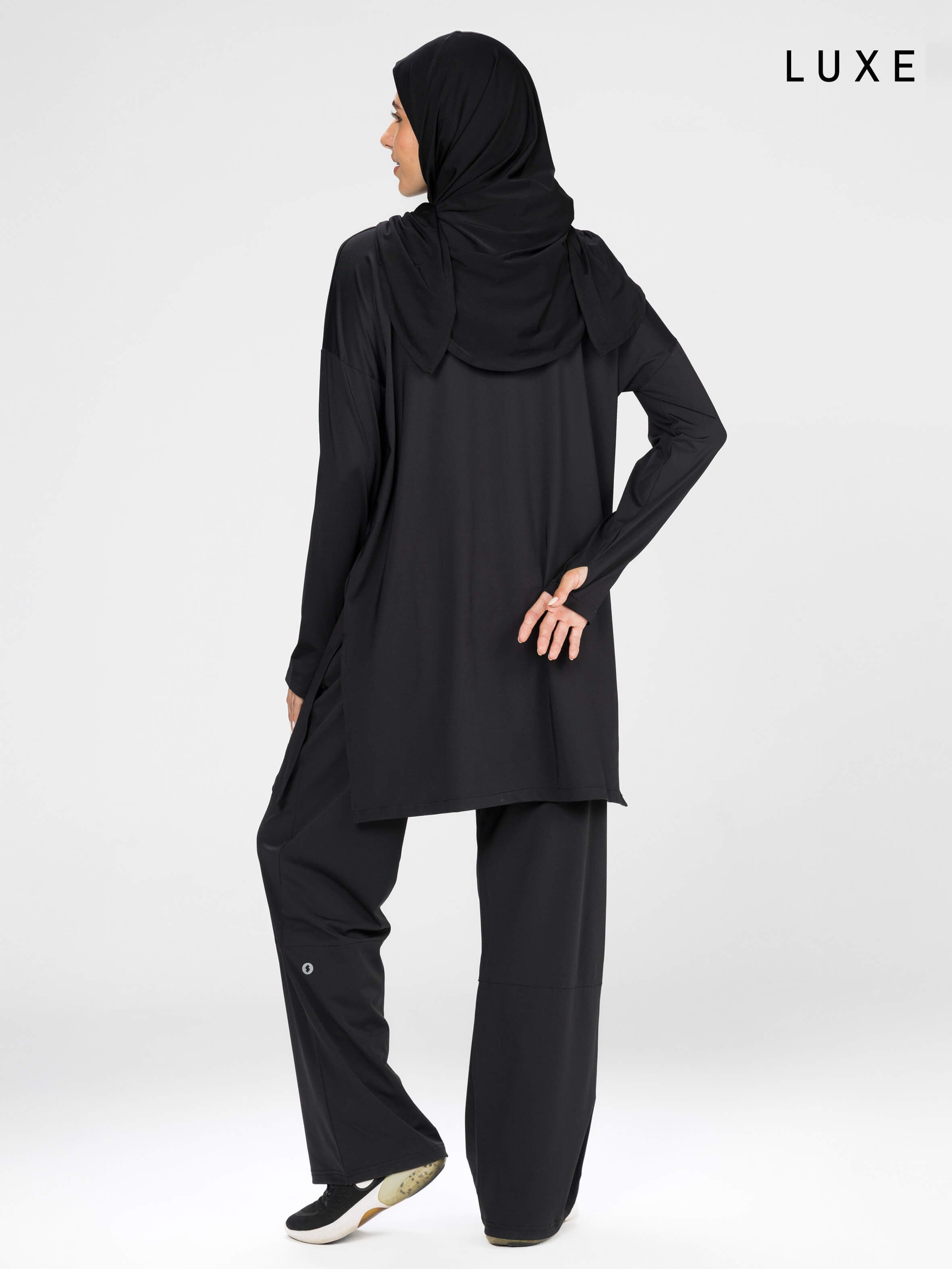 Woman wearing a long-sleeve black modest loose sports activewear top with a hijab on a white background, branded 'LUXE'.