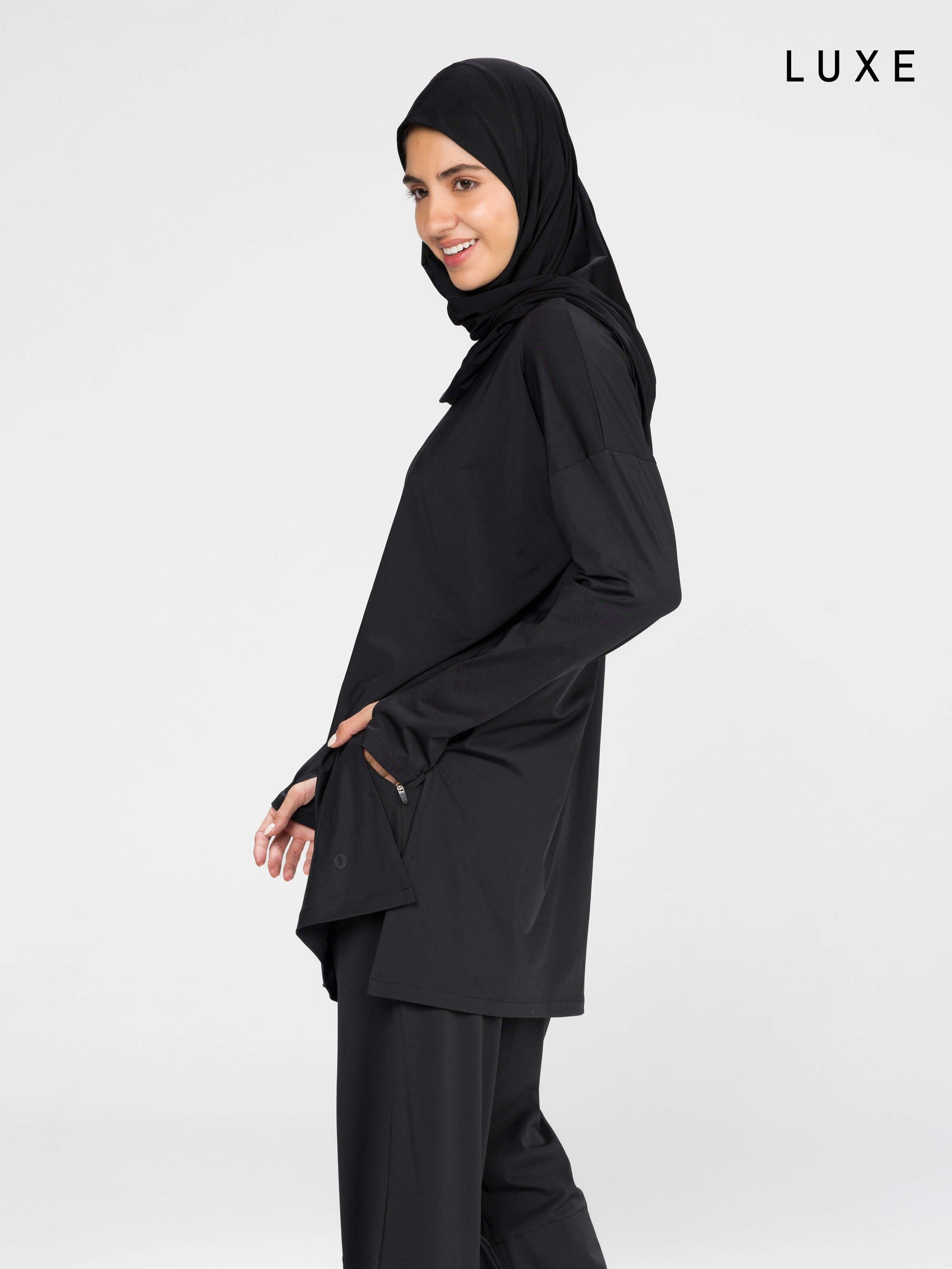 Woman wearing a long-sleeve black modest loose sports activewear top with a hijab on a white background, branded 'LUXE'.